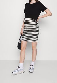 Robe de maternité avec un haut noir et une jupe moulante à rayures noires et blanches, en tissu doux. Le mannequin porte des baskets blanches et tient un sac noir.