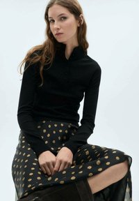 Musta pitkähiha toppi, jossa on henley-kaulus ja nappilista, yhdistettynä polvipituiseen mustaan hameeseen, jossa on kultaisia pilkkuja.