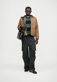 Jachetă maro matlasată cu guler moale, pulover în dungi verde închis și bleumarin dedesubt, pantaloni negri largi și o geantă de umăr neagră.