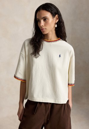 Vrouw met lang donker haar, gekleed in een wit kort mouwig shirt met rode en gele bies en een bruine broek, staand tegen een neutrale achtergrond.