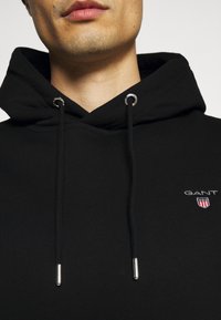 Zwarte hoodie van zacht materiaal met een trekkoord met metalen uiteinden. GANT-logo en embleem in wit en rood op de linkerborst.