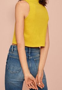 Modelo vistiendo una blusa sin mangas de color amarillo brillante y unos pantalones vaqueros de cintura alta azules, de pie frente a un fondo rosa claro con las manos entrelazadas detrás.