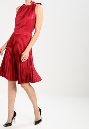 Frau trägt ärmelloses rotes Satinkleid mit Schleife auf der Schulter und plissiertem Rock, kombiniert mit schwarzen High Heels, steht vor weißem Hintergrund.