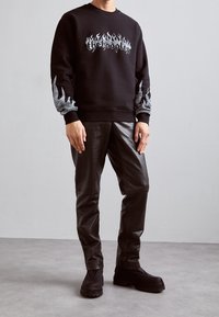 Svart sweatshirt med flamdekor och grafisk text, kombinerad med glänsande svarta fusk läderbyxor och kraftiga svarta ankelstövlar.