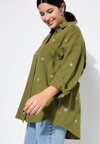 Olijfgroene button-up blouse in een textuurstof, met zilveren sterborduursels en opgerolde mouwen, gecombineerd met lichtblauwe jeans.