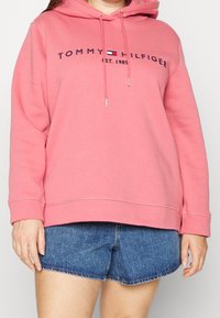 Vaaleanpunainen huppari, joka on valmistettu pehmeästä kankaasta. Edessä logo, jossa lukee "TOMMY HILFIGER" ja "EST. 1985." Kiristysnyörit metallipäillä, rento istuvuus.