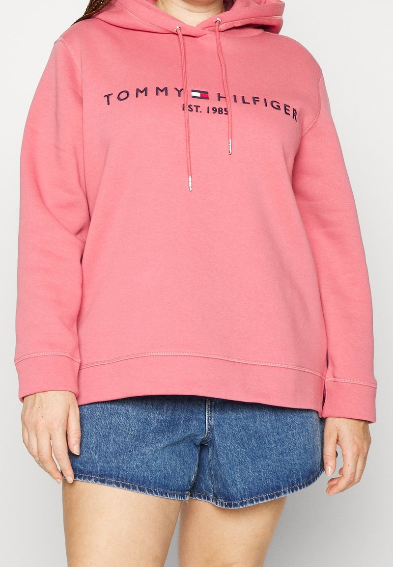 Vaaleanpunainen huppari, joka on valmistettu pehmeästä kankaasta. Edessä logo, jossa lukee "TOMMY HILFIGER" ja "EST. 1985." Kiristysnyörit metallipäillä, rento istuvuus.