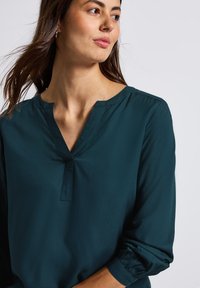 Blusa teal con scollatura a V e maniche lunghe. Realizzata in un tessuto liscio, presenta dettagli plissettati ai polsi e una vestibilità rilassata.