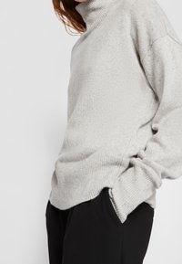 Szary dziergany sweter z golfem, o luźnym kroju, z prążkowanym kołnierzem i fakturowanym materiałem. Połączony z czarnymi spodniami.