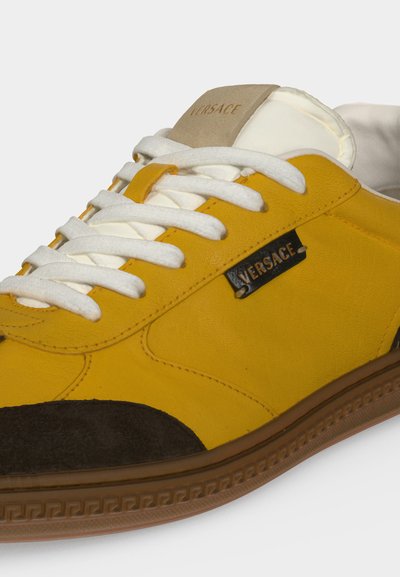 Baskets Versace jaunes et noires avec lacets blancs, étiquette beige sur la languette et semelle marron à motif.
