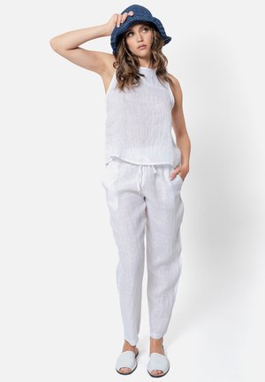 Donna con cappello bucket blu, top bianco senza maniche, pantaloni bianchi con coulisse e sandali bianchi, in piedi con una mano in tasca.