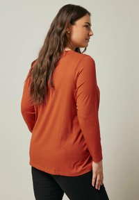 Ribbet langærmet top i brændt orange, med en rund halsudskæring, afslappet pasform og ensfarvet design, parret med sorte bukser.