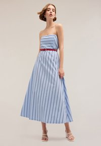Rochie maxi fără bretele din țesătură bleu deschis cu dungi verticale bleumarin, accentuată cu o curea roșie; textură netedă și siluetă fluidă.