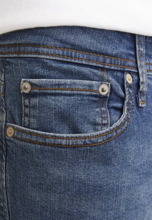 Dark blue denim jeans with visible stitching, a small coin pocket, and metal hardware accents, showing a textured fabric surface.