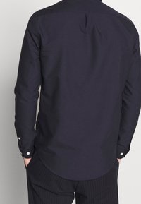Camisa de manga larga de color azul marino con un dobladillo curvado, puños abotonados y una textura suave. Presenta costuras pronunciadas en los hombros.