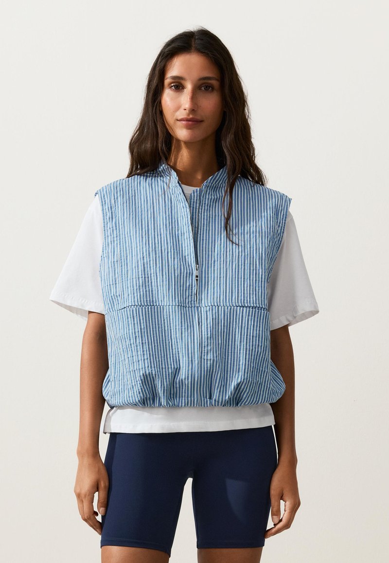 Femme aux longs cheveux foncés portant un gilet sans manches rayé bleu et blanc zippé sur un t-shirt blanc à manches courtes et un short de cyclisme bleu marine.