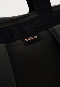 Close-up of dark green and black fabric bag with black handle and small rectangular Barbour logo tag.