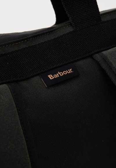 Gros plan sur un sac en tissu noir et vert foncé avec une poignée noire et une petite étiquette rectangulaire au logo Barbour.