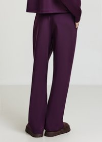 Dunkellila Lounge-Hosen aus glattem Stoff mit entspanntem Schnitt und Seitentaschen, kombiniert mit braunen Schuhen.