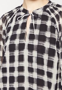 Blouse à carreaux noir et blanc avec un col en clé, détails à cordon de serrage et manches froncées, réalisée dans un tissu léger.