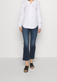 Camisa blanca de manga larga con escote en V, combinada con jeans acampanados de color azul oscuro y sandalias negras tipo slip-on. Enfocarse en la tela y el corte.