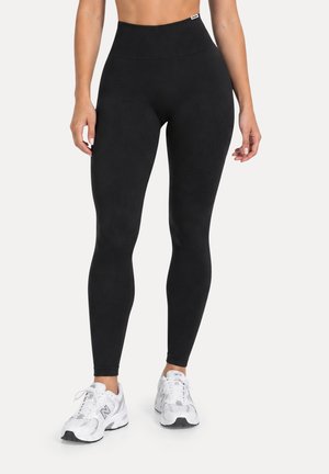 Mujer que lleva puestos unos leggings negros de cintura alta y zapatillas deportivas blancas, de pie con los brazos relajados a los lados sobre un fondo blanco.