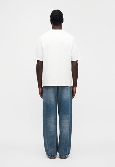 Λευκό κοντομάνικο μπλουζάκι συνδυασμένο με μπλε τζιν παντελόνι φαρδιάς γραμμής. Το μπλουζάκι έχει στρογγυλή λαιμόκοψη· το τζιν διαθέτει κλασικό σχέδιο με πέντε τσέπες και ξεθωριασμένες λεπτομέρειες.