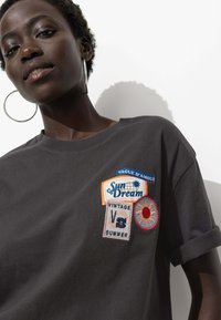 T-shirt en coton gris anthracite avec des manches retroussées, ornée de patchs brodés multicolores étiquetés "Sun Dream", "Vintage" et "Summer".