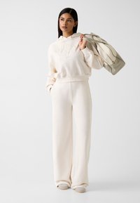 Roomkleurige hoodie met reliëflogo, gecombineerd met bijpassende wijde broekspijpen. Model houdt een lichtgrijze handtas met dubbele handvatten vast.