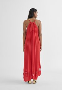 Červené plisované maxi šaty s halter krkom, detailom mašle a zlatými akcentmi na ramenách; obsahujú čipkovaný lem na spodku.