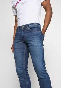 Blå denimjeans med slim fit, med fem fickor, subtila blekningar och en grön knapppunkt på midjan.