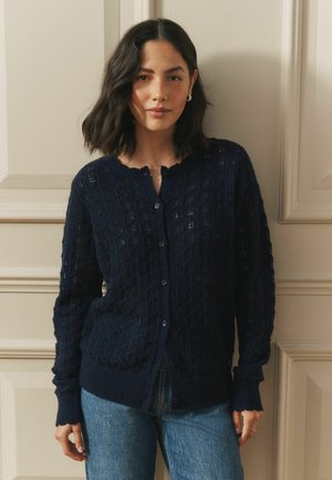 Frau mit schulterlangem dunklem Haar trägt eine dunkelblaue, strukturierte Strickjacke und blaue Jeans, steht in Innenräumen vor einer beigen, vertäfelten Wand.