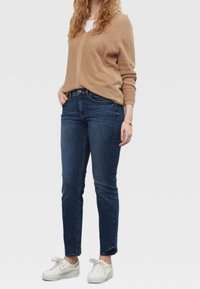 Mujer vestida con un suéter beige, jeans azul oscuro y zapatillas blancas, de pie con una mano en el bolsillo frente a un fondo liso.