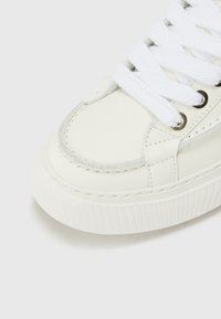 Baskets en cuir blanc avec bout rond, semelle texturée et lacets plats blancs maintenus par des œillets en acier inoxydable. Présente un détail de couture sur le dessus.