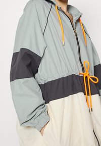 Chaqueta ligera en bloques de color con un diseño en gris, negro y crema. Equipado con una cinturilla elástica y detalles en naranja en los cordones.
