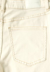 Tejido de mezclilla blanca con costuras claras. Presenta un solo bolsillo trasero con una ligera curva y una etiqueta en la cinturilla que dice "Street One Studio."