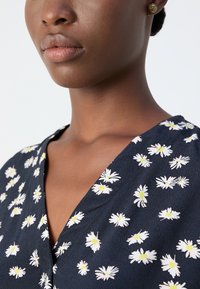 Blusa floreale blu navy con scollo a V, decorata da motivi di margherite bianche con centri gialli e tessuto testurizzato. Orecchini a bottone dorati visibili.