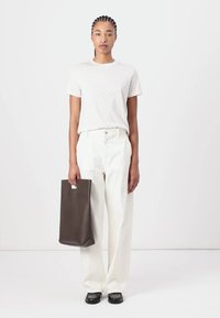 White textured t-shirt, loose cream trousers, and black loafers. Holding a dark brown tote bag with a cut-out handle. Simple design.