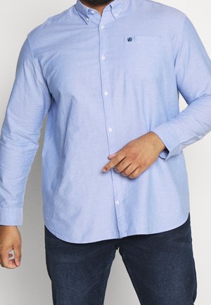 Mand i lyseblå skjorte med knapper, peger nedad, iført mørke jeans, foran en ensfarvet lys baggrund.