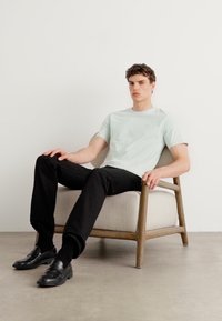 T-shirt de manga curta verde-claro, calças pretas, sentado numa cadeira de madeira minimalista com um cushion bege, contra um fundo branco liso.