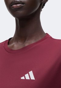 Gros plan d’une personne portant un maillot de sport rouge foncé avec un petit logo blanc Adidas sur la poitrine.
