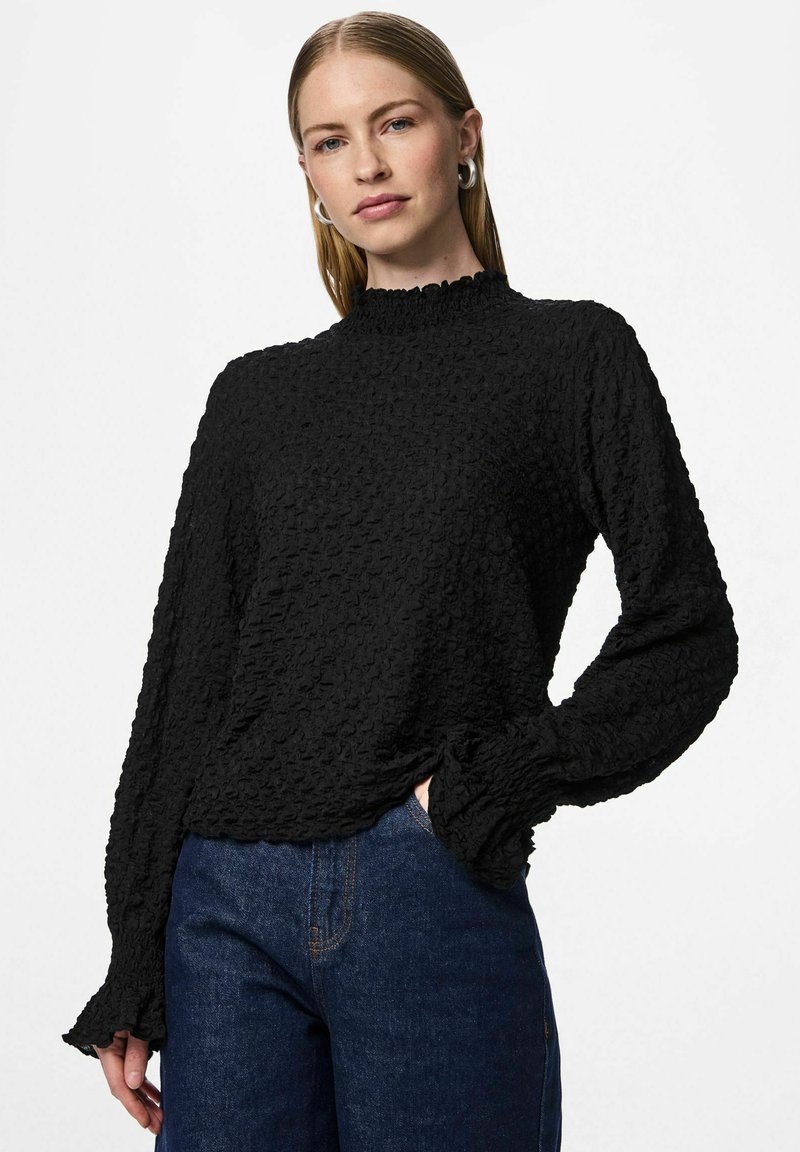 Crni teksturirani dugi rukav s visokim naboranim ovratnikom i raširenim manšetama, nošen s plavim trapericama. Tkanina ima naborani uzorak.