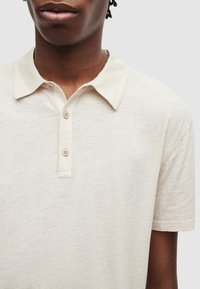 Polo beige à texture douce, avec un col pointu, une patte de boutonnage à trois boutons et des manches courtes. Tissu légèrement chiné.