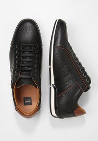 Baskets en cuir noir avec surface texturée, accents marron contrastants, cinq paires d'œillets et semelle en caoutchouc blanche.