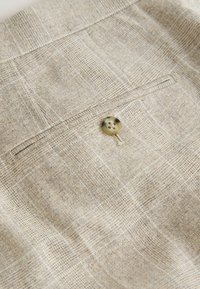 Beiger strukturierter Stoff mit einem dezenten Karomuster, der eine mit Knöpfen versehene Gesäßtasche an maßgeschneiderten Hosen zeigt.