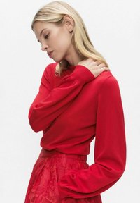 Czerwona, długorękawowa sweter wykonany z miękkiej dzianiny, z okrągłym dekoltem i luźnym krojem. Noszony z czerwoną warstwowaną spódnicą.