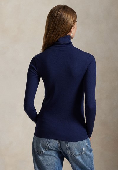 Kobieta w granatowym, długim swetrze z golfem i jasnoniebieskich postrzępionych dżinsach, stojąca na neutralnym tle.