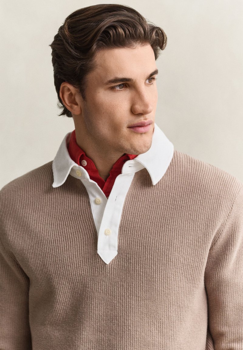 Young man with styled brown hair wearing a beige sweater over a white collared shirt and red undershirt, looking to the side against a plain background.