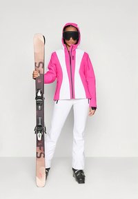 Γυναίκα με ροζ και άσπρη σαμπρέλα σκι, άσπρα παντελόνια, μαύρες μπότες σκι και γυαλιά κατά του ήλιου, κρατώντας έναν μόνο κατακόρυφο σκι σε έναν απλό φόντο.