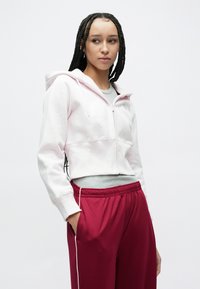 Trumpas rožinis džemperis su priekiu užtrauktuku ir raišteliu, derinamas su raudonomis sportinėmis kelnėmis su šoninėmis juostelėmis ir elastinga juosmens juosta.